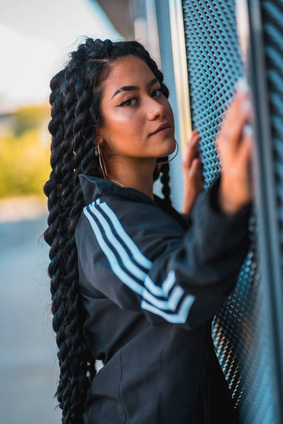 Urban session. Young black ethnic woman with long braids and with tattoos, looking glass, smiling - Foto, Bild