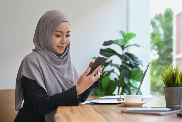 Başörtüsü takan Müslüman iş kadını dizüstü bilgisayarın önünde oturuyor ve cep telefonu kullanıyor.. - Fotoğraf, Görsel