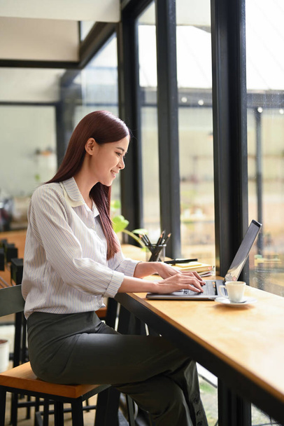 Yan bakış genç serbest çalışan kadın kahve dükkanında ahşap tezgahta oturuyor ve dizüstü bilgisayar kullanıyor.. - Fotoğraf, Görsel