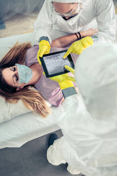 Вид зверху на лікарів у захисному костюмі, які вивчають рентген хворого на планшеті
 - Фото, зображення