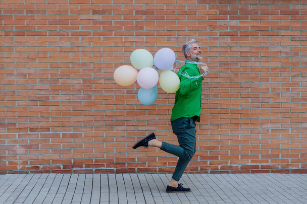 A fun portrait of happy energetic mature man holding balloons and running in street , feeling free. - Foto, Bild