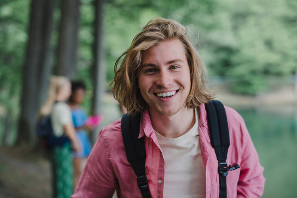 A portrait of young man with friends at background on a hiking or camping trip in the mountains in summer. - Фото, изображение
