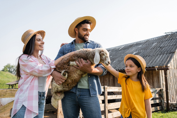 happy farmer in straw hat holding lamb near daughter and wife on farm - Photo, image