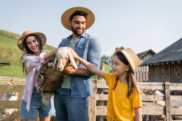 woman and child in straw hats stroking lamb in hands of smiling farmer  - Foto, imagen