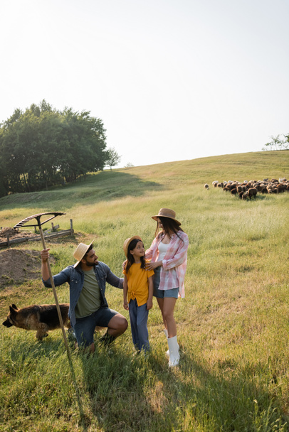 farmář s hráběmi v blízkosti dcery a manželky stojící na pastvinách v blízkosti pasoucího se stáda - Fotografie, Obrázek