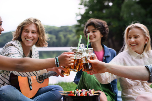 Um grupo de jovens amigos multirraciais acampando perto do lago, tendo churrasco e brinde com bebidas de coquetel. - Foto, Imagem