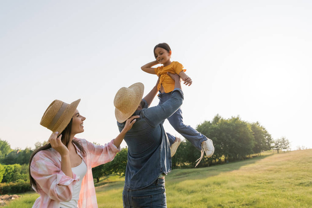 man in straw hat raising up cheerful daughter near happy wife  - Foto, immagini