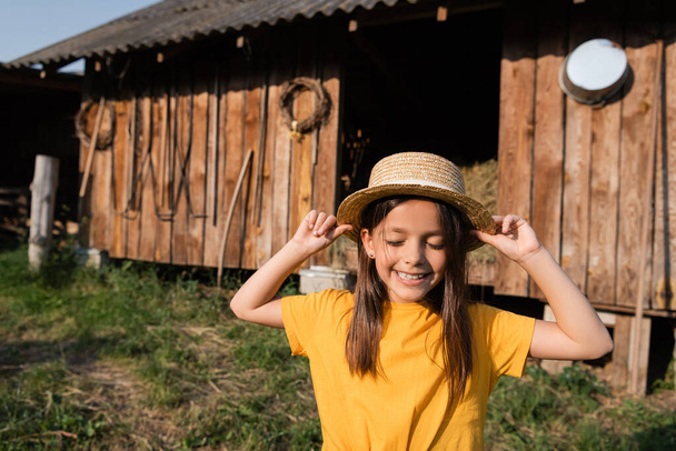 smiling child with closed eyes touching straw hat near wooden barn on blurred background - Foto, imagen