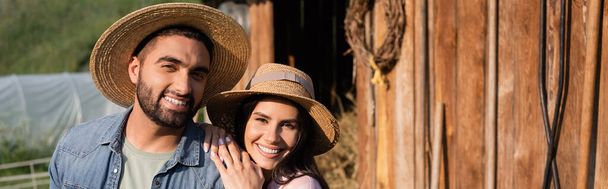 χαρούμενο ζευγάρι αγροτών με ψάθινα καπέλα χαμογελώντας στην κάμερα στην οικογενειακή φάρμα, πανό - Φωτογραφία, εικόνα