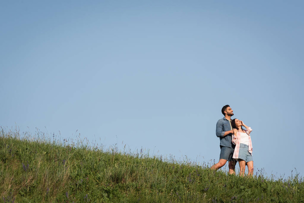 boldog pár farmernadrágban áll a zöld dombon a kék ég alatt, és félrenéz - Fotó, kép