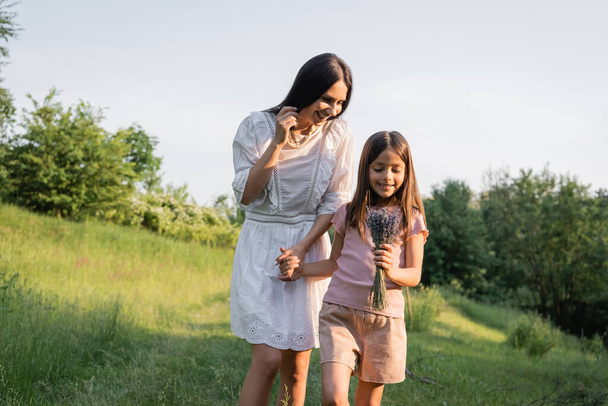 smiling girl with bouquet of lavender holding hands with pleased mom in countryside - Fotografie, Obrázek