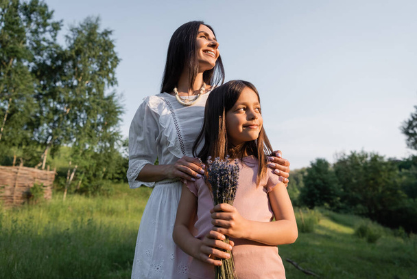 happy woman hugging shoulders of daughter holding lavender flowers outdoors - 写真・画像