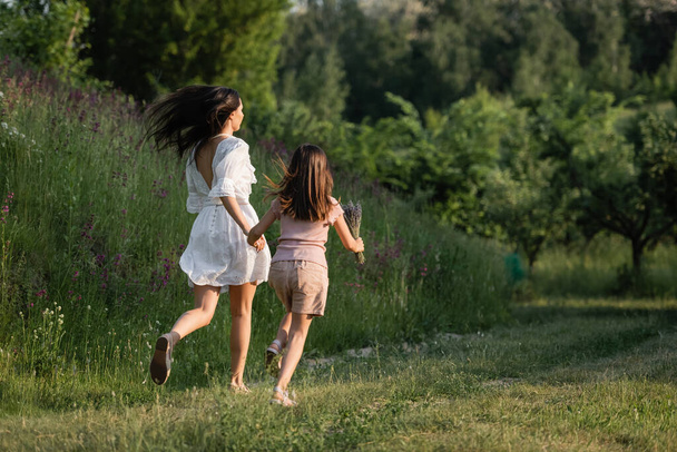 Rückansicht von Mutter und Kind, die sich an Händen halten und auf Graspfad in Richtung Wald laufen - Foto, Bild