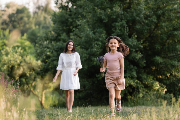 full length of cheerful girl with lavender bouquet running near mom on blurred background - Photo, image