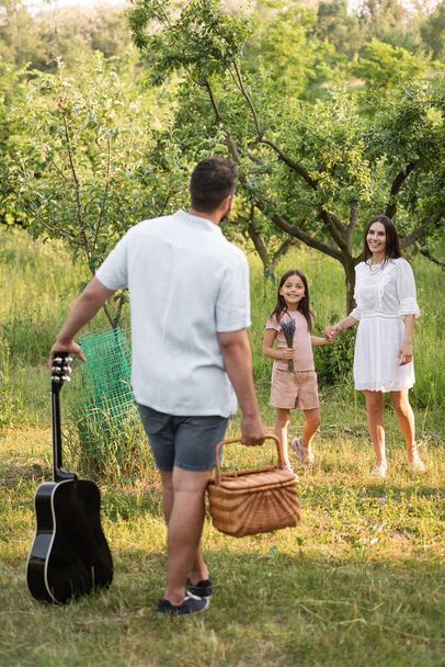man with wicker basket and acoustic guitar near wife and daughter smiling in garden - Foto, afbeelding