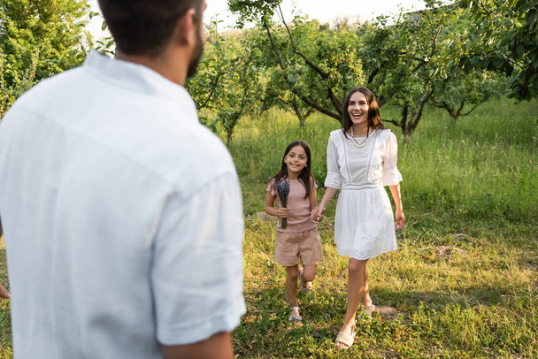  blurred man near happy wife and daughter holding hands near green trees - Valokuva, kuva