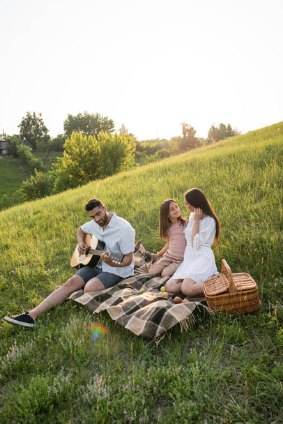 man playing guitar near family sitting on plaid blanket in green meadow - Photo, Image