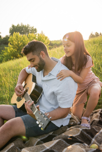 smiling girl hugging shoulders of dad playing guitar while resting in countryside - Zdjęcie, obraz