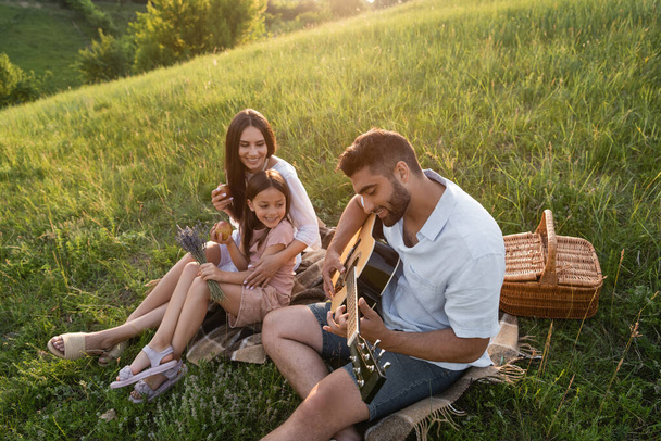 man playing acoustic guitar to happy wife and daughter in green hill on summer day - Foto, Imagem