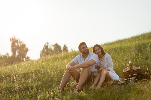 joyful couple sitting on green slope near wicker basket and looking away  - Fotó, kép
