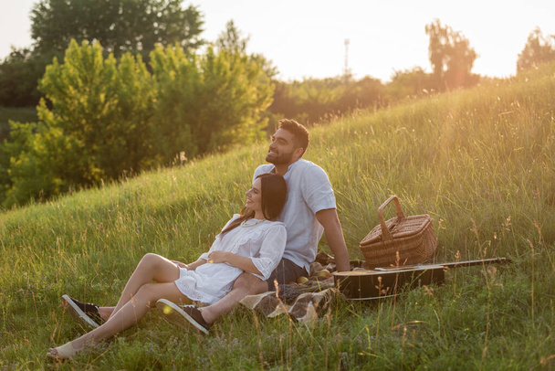 cheerful couple looking away while sitting on green slope near wicker basket and acoustic guitar - Foto, Imagem