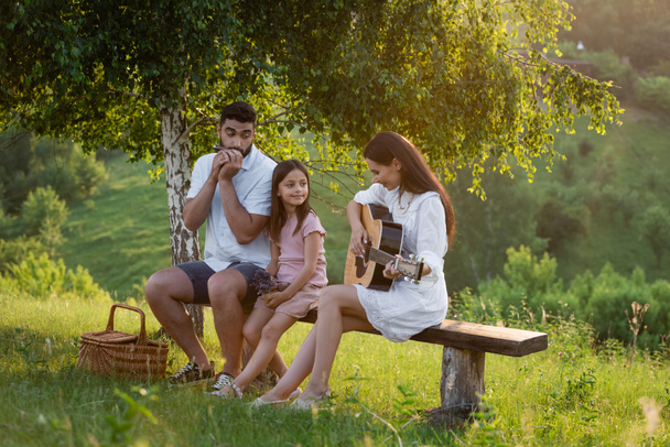 пара играет на губной гармошке и гитаре дочери на скамейке под березой - Фото, изображение