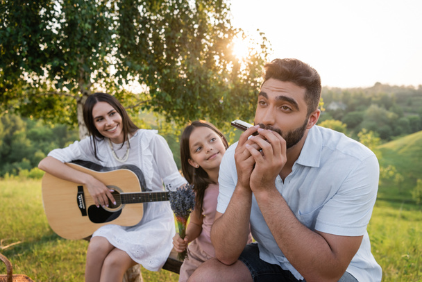 άνδρας παίζει φυσαρμόνικα κοντά κόρη με μπουκέτο και γυναίκα με ακουστική κιθάρα - Φωτογραφία, εικόνα