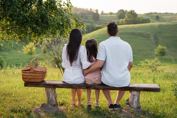 vue arrière de la famille assise sur un banc près du panier en osier devant sur un paysage pittoresque - Photo, image