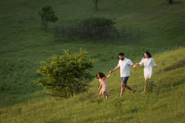 cheerful family holding hands while running on grassy slope on summer day - 写真・画像