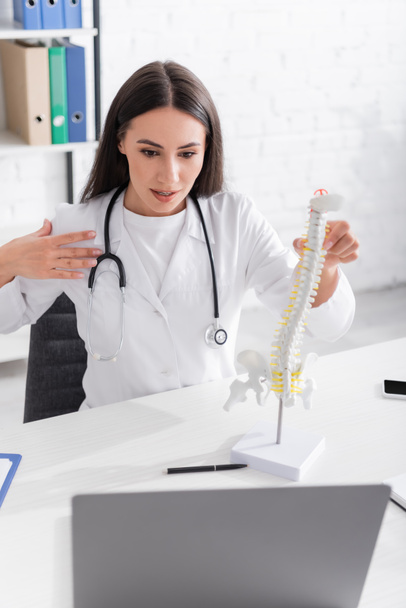 Brunette doctor holding spinal model during online consultation on laptop in hospital  - Photo, image