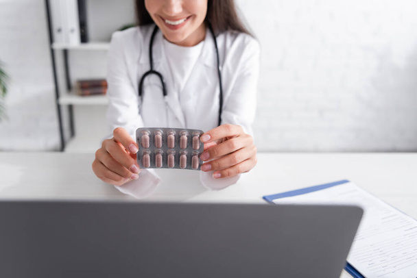 Cropped view of smiling doctor holding blister with pills near laptop and clipboard in clinic  - 写真・画像