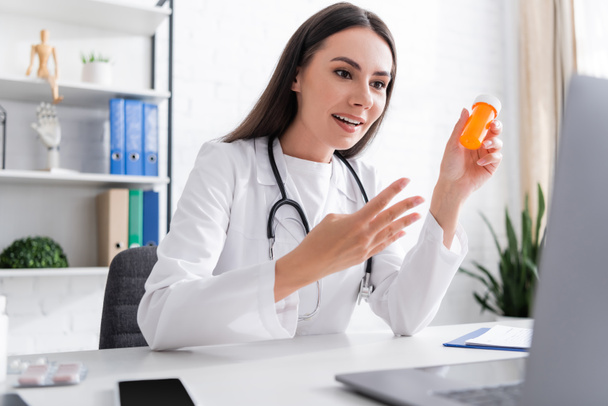 Χαμογελώντας γιατρός κρατώντας χάπια και gesturing κατά τη διάρκεια της κλήσης βίντεο στο φορητό υπολογιστή στην κλινική  - Φωτογραφία, εικόνα