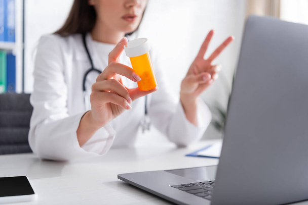 Cropped view of blurred doctor holding jar with pills near gadgets in clinic  - Foto, imagen