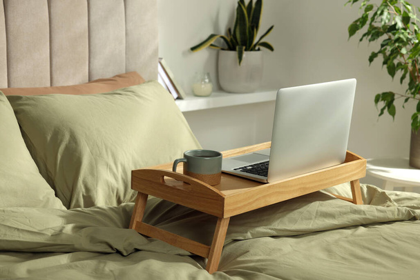 Wooden tray with modern laptop and cup of aromatic tea on bed indoors - Фото, зображення