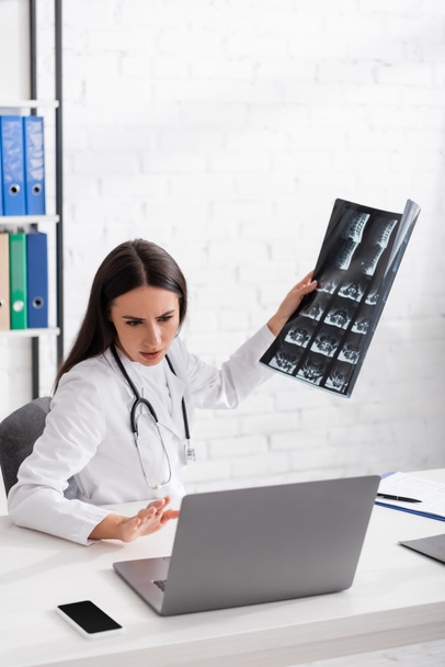 Médico sosteniendo mri scan durante la consulta en línea en el ordenador portátil en la clínica  - Foto, imagen