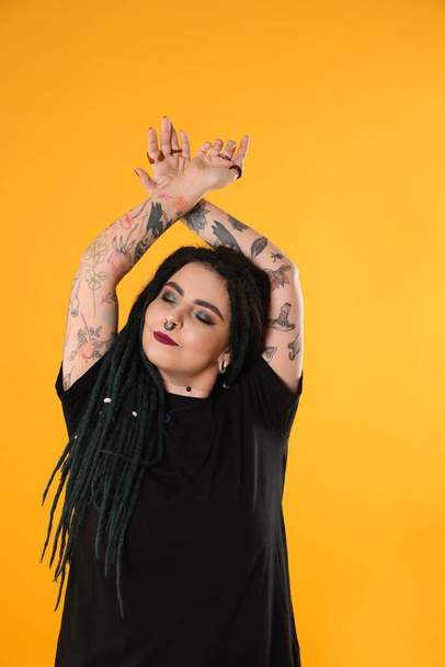 Beautiful young woman with tattoos on arms, nose piercing and dreadlocks against yellow background - Foto, imagen