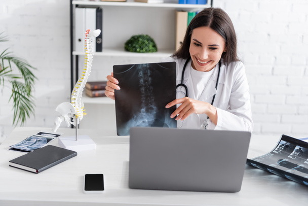 Positive doctor holding x-ray scan during online consultation on laptop in hospital  - Фото, изображение