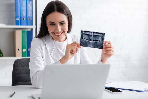 Positive doctor holding scan of teeth during video chat on laptop in hospital  - Valokuva, kuva