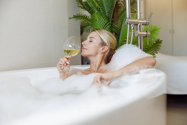 Side view on young beautiful Caucasian female enjoying white wine and taking bath. Bathroom. Resting - Fotografie, Obrázek