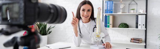 Smiling doctor gesturing near spinal model and blurred digital camera in clinic, banner  - Fotoğraf, Görsel