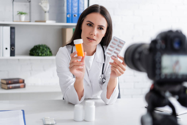 Doctor in white coat holding pills near blurred digital camera in clinic  - Foto, afbeelding