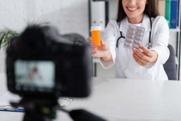 Vue recadrée d'un médecin souriant tenant des pilules près d'un appareil photo numérique flou dans une clinique  - Photo, image