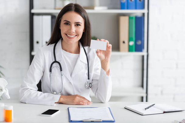 Smiling doctor holding business card with copy space near smartphone and clipboard in clinic  - Fotoğraf, Görsel