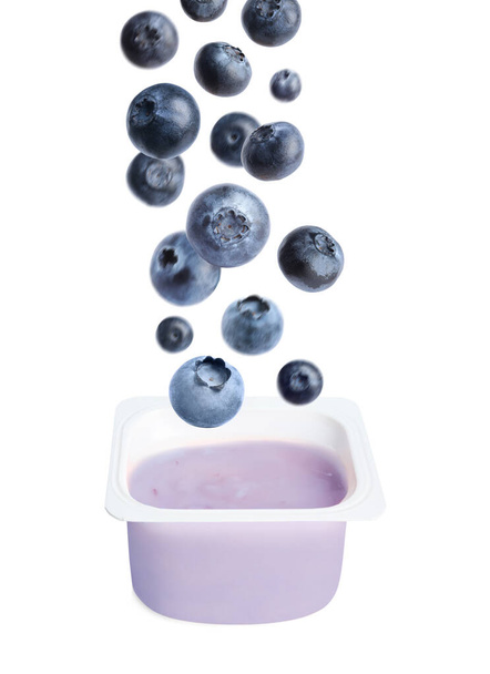 Many fresh blueberries falling into plastic cup with yogurt on white background - Foto, imagen