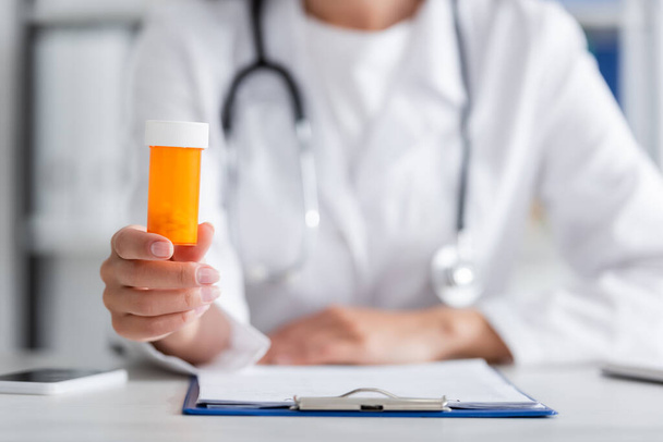 Cropped view of blurred doctor holding pills near clipboard and smartphone in clinic  - Photo, Image