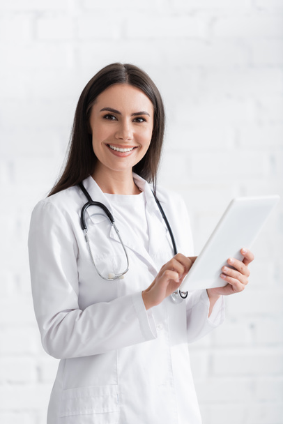 Brunette doctor smiling at camera and using digital tablet in clinic  - Фото, изображение