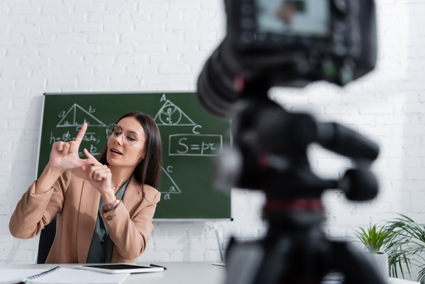 Nauczyciel w okularach gestykulujący w pobliżu tablicy z matematycznymi wzorami i rozmytym aparatem cyfrowym w klasie  - Zdjęcie, obraz