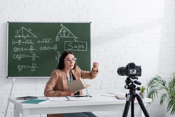 Tutor holding book near digital camera and chalkboard with mathematic formulas in class  - Foto, Imagen