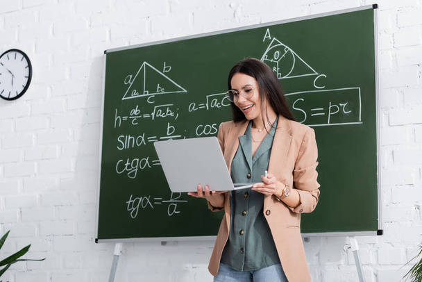 Positive teacher holding laptop during online lecture near chalkboard with math formulas  - 写真・画像