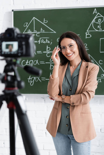 Cheerful teacher holding chalk near chalkboard and digital camera in classroom  - Photo, image